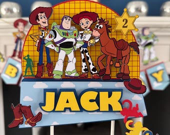 Può includere: Decorazione colorata per torta di compleanno con i personaggi di Toy Story: Woody, Buzz Lightyear, Jessie e un cavallo marrone. Il nome "JACK" è in lettere gialle, con un distintivo "2". Stivali da cowboy e scimmie decorano la torta.