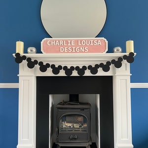May include: A white fireplace mantel with a black wood-burning stove. A black and white Mickey Mouse garland hangs above the mantel with a pink sign that reads "Charlie Louisa Designs".