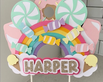 Puede incluir: Un adorno para pastel de arcoíris con temática de dona, caramelo y helado. El adorno presenta un arcoíris con piezas de caramelo, una dona y conos de helado. El nombre "HARPER" está escrito en letras doradas con purpurina sobre un fondo rosa.