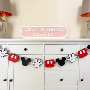 May include: A Mickey Mouse themed party banner featuring red shorts, white gloves, and black Mickey Mouse heads. The banner is hanging on a white dresser with a sign that reads "Charlie Louisa Designs".