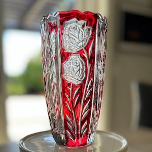 May include: A tall, clear glass vase with a vibrant red interior. The vase features etched floral designs and vertical leaf patterns. The vase is displayed on a small, white, round pedestal.