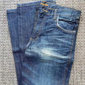 K&ouml;nnte beinhalten: Eine blaue Jeans aus Denim, die in der H&auml;lfte gefaltet ist. Die Jeans hat eine helle Waschung und einen verblassten Look. Das Wrangler-Logo ist auf der Ges&auml;&szlig;tasche sichtbar.