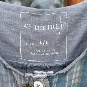 May include: A gray and blue plaid shirt with a button closure and a star-shaped button. The shirt has a label that reads "We The Free" and "Size L/G" and "Made in China, Fabrique en Chine".