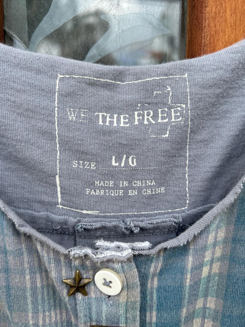 May include: A gray and blue plaid shirt with a button closure and a star-shaped button. The shirt has a label that reads "We The Free" and "Size L/G" and "Made in China, Fabrique en Chine".
