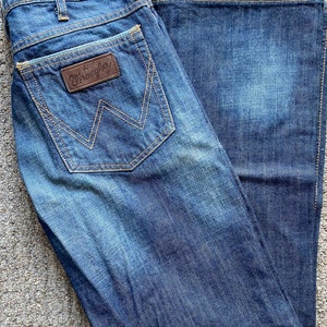 K&ouml;nnte beinhalten: Eine blaue Jeanshose mit einem braunen Leder-Patch auf der Ges&auml;&szlig;tasche, auf dem "Wrangler" steht.