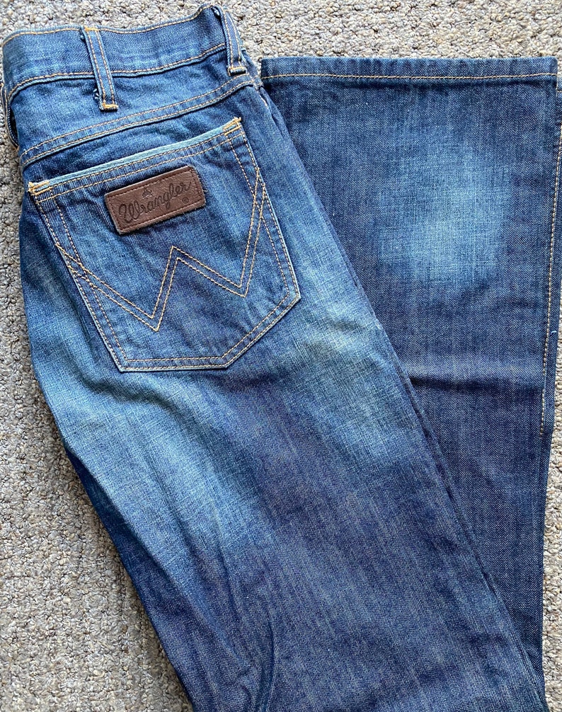 K&ouml;nnte beinhalten: Eine blaue Jeanshose mit einem braunen Leder-Patch auf der Ges&auml;&szlig;tasche, auf dem "Wrangler" steht.