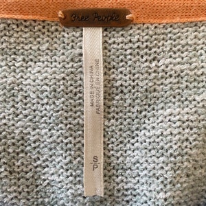 May include: A gray and blue striped sweater with a Free People tag. The tag is attached to a white fabric label that reads "Made in China, Fabriqu&eacute; en Chine, T/C 100".