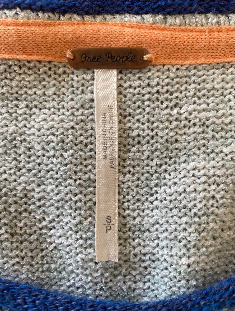 May include: A gray and blue striped sweater with a Free People tag. The tag is attached to a white fabric label that reads "Made in China, Fabriqu&eacute; en Chine, T/C 100".