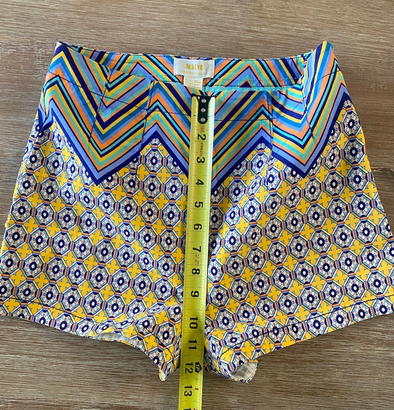 May include: Yellow and blue patterned shorts with a zig-zag pattern at the top and a smaller floral pattern on the bottom. The shorts have a waistband with a label that reads "Malva".