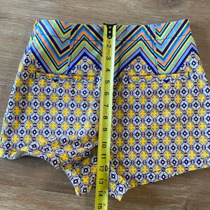 May include: Yellow, blue, and white patterned shorts with a zig-zag pattern at the waistband. The shorts are measured with a tape measure.