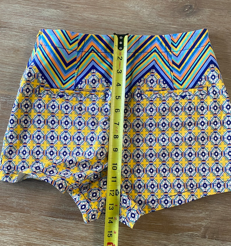 May include: Yellow, blue, and white patterned shorts with a zig-zag pattern at the waistband. The shorts are measured with a tape measure.