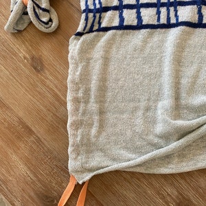 May include: A gray knitted sweater with a blue and white geometric pattern. The sweater has a drawstring at the bottom with orange and yellow cords.