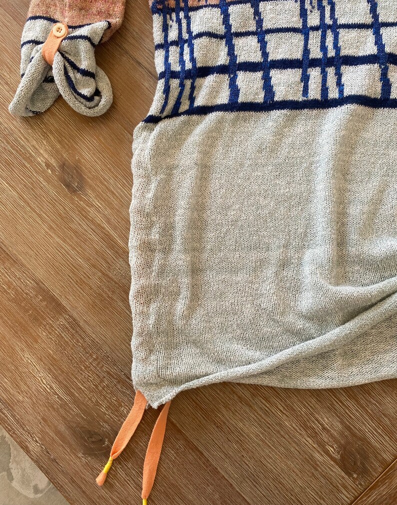 May include: A gray knitted sweater with a blue and white geometric pattern. The sweater has a drawstring at the bottom with orange and yellow cords.
