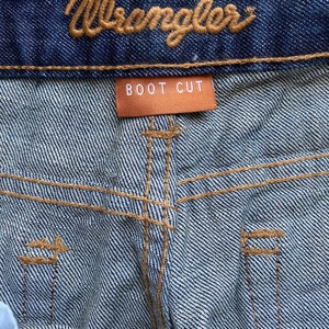 K&ouml;nnte beinhalten: Nahaufnahme einer blauen Jeanshose mit einem braunen Leder-Wrangler-Etikett und einem braunen Etikett mit der Aufschrift "BOOT CUT".