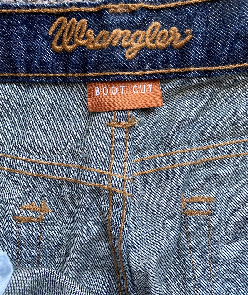 K&ouml;nnte beinhalten: Nahaufnahme einer blauen Jeanshose mit einem braunen Leder-Wrangler-Etikett und einem braunen Etikett mit der Aufschrift "BOOT CUT".