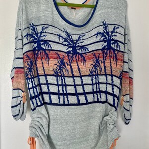 May include: A light gray sweater with a palm tree print and blue and pink stripes. The sweater has a scoop neck and long sleeves with buttons on the cuffs. The bottom of the sweater has a drawstring.