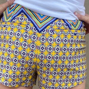 May include: Yellow and blue patterned shorts with a geometric design. The shorts have a waistband with a blue, green, and orange striped pattern.