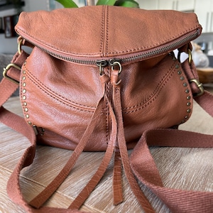 May include: A brown leather crossbody bag with a zippered top and adjustable strap. The bag features decorative stitching, metal studs, and a tassel detail. The bag is made of a soft, textured leather.