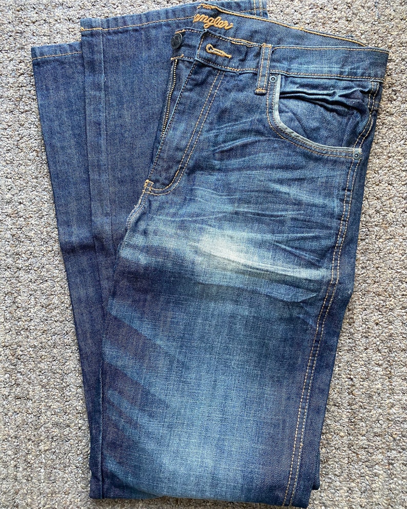 K&ouml;nnte beinhalten: Eine blaue Jeans aus Denim, die in der H&auml;lfte gefaltet ist. Die Jeans hat eine helle Waschung und einen verblassten Look. Das Wrangler-Logo ist auf der Ges&auml;&szlig;tasche sichtbar.