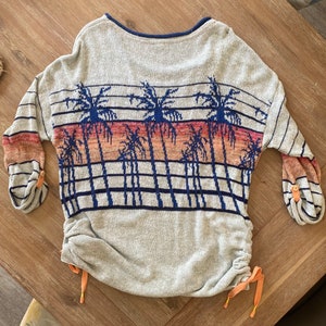 May include: A gray sweater with a palm tree print and a drawstring at the bottom. The sweater has a blue and orange color scheme.