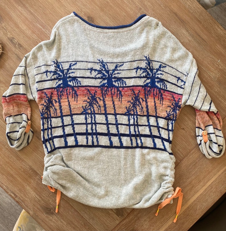 May include: A gray sweater with a palm tree print and a drawstring at the bottom. The sweater has a blue and orange color scheme.