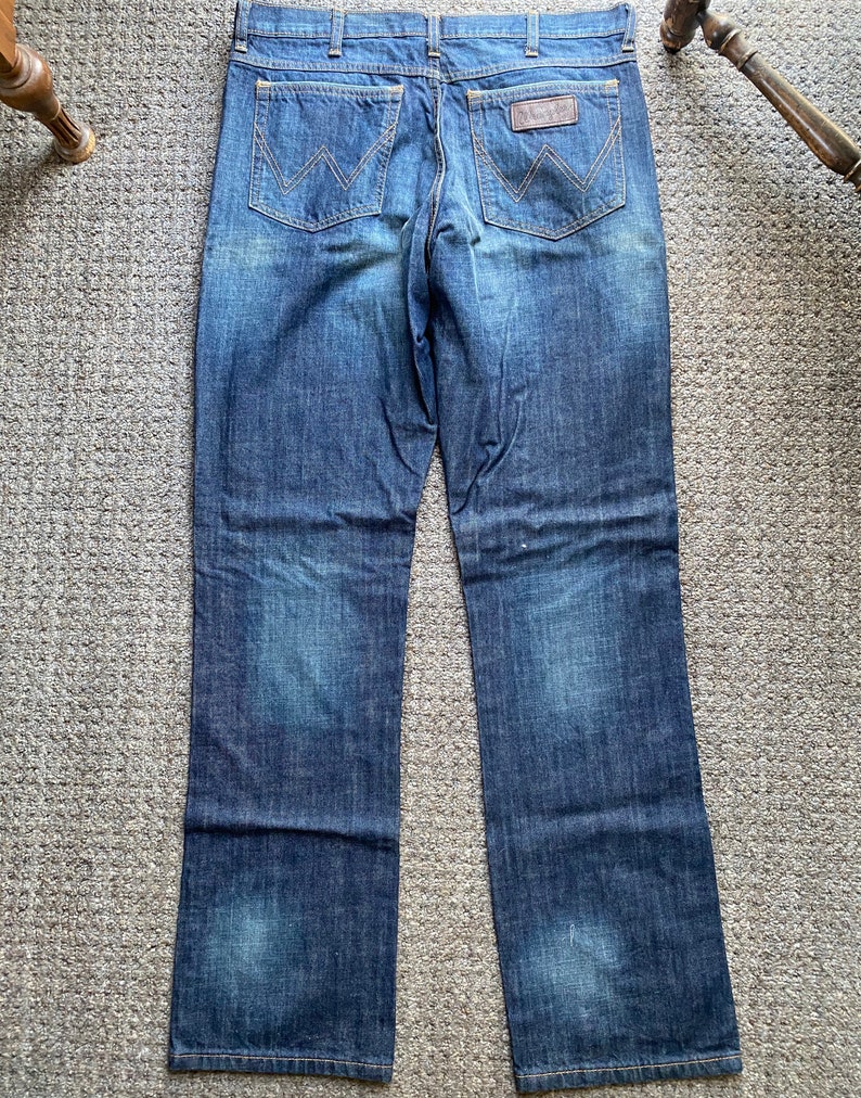 K&ouml;nnte beinhalten: Eine dunkelblaue Jeanshose mit einem braunen Leder-Patch auf der Ges&auml;&szlig;tasche, auf dem "Wrangler" steht.