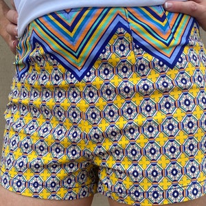 May include: Yellow and blue patterned shorts with a geometric design. The shorts have a waistband with a blue, orange, and yellow striped pattern.