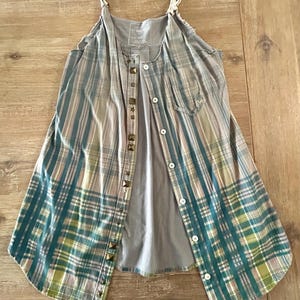 Puede incluir: Un vestido de camisa de cuadros sin mangas con un top de tirantes gris debajo. El vestido tiene un frente abotonado y está decorado con tachuelas metálicas.