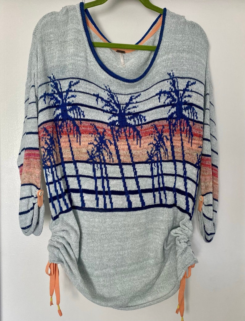 May include: A light blue sweater with a palm tree print and horizontal stripes. The sweater has a scoop neck and long sleeves with buttons on the cuffs. The bottom of the sweater has a drawstring with orange ties.