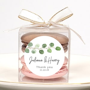 May include: A clear box with a white ribbon tied around it. Inside the box are two macarons, one pink and one brown. A round sticker with a green leafy branch design and the text "Juliana & Harry Thank you 11.22.29" is on the front of the box.