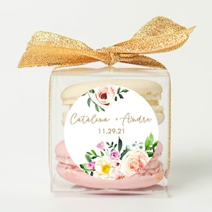 May include: A clear box with a gold ribbon tied around it. Inside the box are three macarons, one white and two pink. A round sticker with a floral design and the text "Catalina + Andre 11.29.21" is on the front of the box.