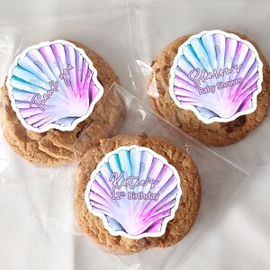 May include: Three clear plastic bags each containing a chocolate chip cookie with a round sticker on top. The stickers feature a watercolor seashell design with different text on each. One sticker says "Thank you", another says "Sharon's Baby Shower", and the last one says "Katie's 12th Birthday".