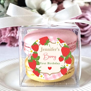 May include: A clear, square box tied with a white ribbon, containing two macarons. The top macaron is pink, the bottom is yellow. A circular sticker with strawberries and the text "Jennifer's Berry First Birthday" is on the box.