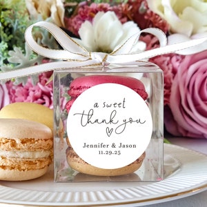 May include: A clear box filled with pastel-colored macarons, tied with a white ribbon with gold trim. A white round sticker with black text reads "a sweet thank you" with a heart symbol and the names "Jennifer & Jason" and the date "11.29.25".