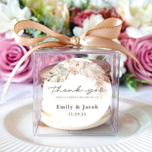 May include: A clear plastic box with a gold ribbon tied around it. Inside the box are three macarons stacked on top of each other. The macarons are decorated with a round sticker that says "Thank you for celebrating with us Emily & Jacob 11.29.25". The sticker has a floral design with brown and white feathers.