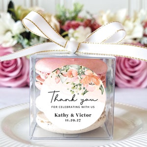 May include: A clear box with a white ribbon tied around it. Inside the box are two pink macarons with a round sticker on top. The sticker has a floral design with peach and pink flowers and green leaves. The text on the sticker says "Thank you for celebrating with us Kathy & Victor 11.29.27".