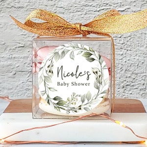 May include: A clear box with a gold ribbon tied around it. Inside the box are macarons with a white and green floral design sticker that says "Nicole's Baby Shower".