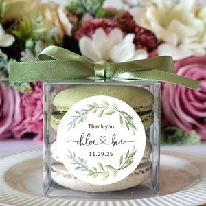 May include: A clear box filled with green macarons, decorated with a round sticker that says "Thank you Chloe & Ben 11.29.25" with a green leafy wreath design.