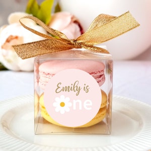 May include: A clear plastic box with a gold ribbon tied around it. Inside the box are two macarons, one pink and one yellow. The macarons have a pink and white sticker on them that says "Emily is one" with a yellow daisy.