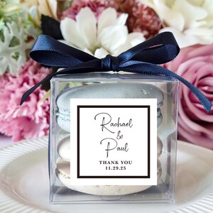 May include: A clear box with a navy blue ribbon tied around it. Inside the box are two macarons, one light blue and one white. A white label with black text reads "Rachael & Paul Thank You 11.29.25".