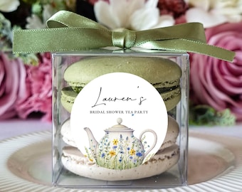 May include: A clear square plastic box containing two stacked macarons, one light green and one off-white, topped with a sage green satin ribbon. A circular sticker on the front features a teapot illustration with wildflowers and the text Lauren's BRIDAL SHOWER TEA PARTY.