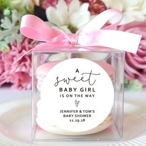May include: A clear box with a pink ribbon tied around it. Inside the box are pink and white macarons with a round white sticker that says "A sweet baby girl is on the way" with a heart and "Jennifer & Tom's Baby Shower 11.29.28"