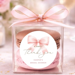May include: A clear box tied with a pink ribbon, containing two macarons. One is chocolate-colored, the other is pink. A circular sticker on the box reads "A Sweet Thank You" and "Harper's Bridal Shower."