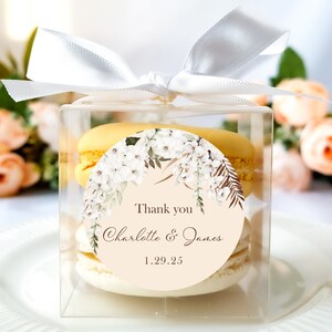 May include: A clear box with a white ribbon tied around it. Inside the box are three yellow macarons with a round sticker on top. The sticker has a floral design and the text "Thank you Charlotte & James 1.29.25".