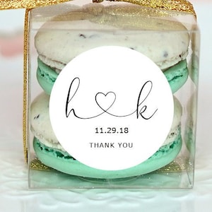 May include: Two mint green macarons with a white circle sticker on top. The sticker has the letters "h" and "k" connected by a heart, the date "11.29.18", and the words "Thank You". The macarons are in a clear plastic box with a gold ribbon.