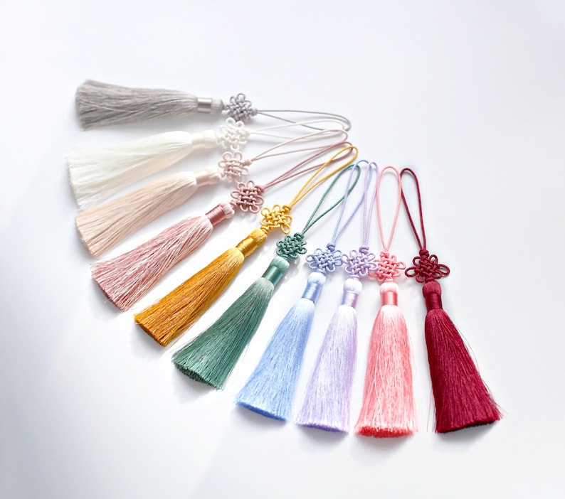 K&ouml;nnte beinhalten: Eine Ansammlung von zehn dekorativen Quasten, die f&auml;cherf&ouml;rmig angeordnet sind. Jede Quaste besitzt seidige Fransen in den Farben Grau, Wei&szlig;, Creme, Rosa, Gold, Gr&uuml;n, Hellblau, Lavendel, Koralle und Dunkelrot, gekr&ouml;nt von einem Zierknoten.
