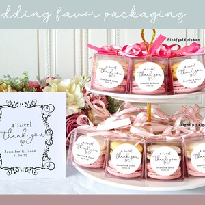 May include: Wedding favor packaging with macarons in clear boxes tied with pink and light pink ribbons. The boxes have a round sticker with the text "a sweet thank you" and the couple's names and wedding date.