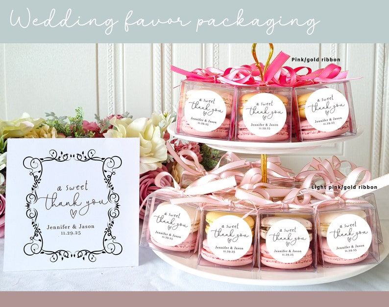 May include: Wedding favor packaging with macarons in clear boxes tied with pink and light pink ribbons. The boxes have a round sticker with the text "a sweet thank you" and the couple's names and wedding date.