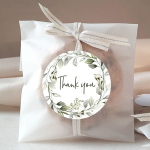 May include: A white paper bag with a brown ribbon tied around it. The bag has a round sticker with a green leafy wreath and the text "Thank you".