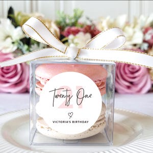 May include: A clear square box tied with a white ribbon with gold trim, containing two macarons. A round sticker on the box reads "Twenty One" with a heart and "Victoria's Birthday". The macarons are pink and beige.